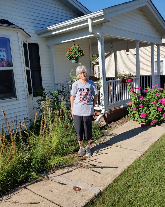 Jan S. standing on the front walk of her Saddlebrook Farms home, wearing a shirt reading "Say no to the T5 data center", with a matching sign in the front window and native plantings along the walk